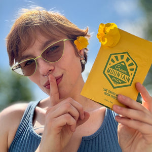 Best Cannabis Dispensary near me person holding a Green Mountain High Lime Ginger Mint Rosin Drops 50mg with a yellow flower in sunglasses, outdoors.